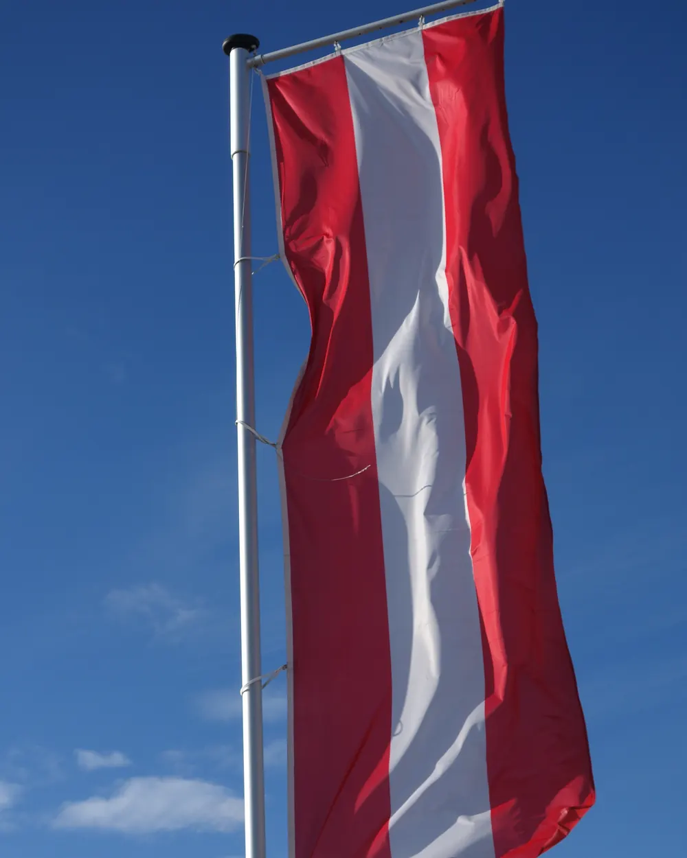 Rot-weiß-rote Fahne im Hochformat an einem Mast mit Ausleger vor blauem Himmel