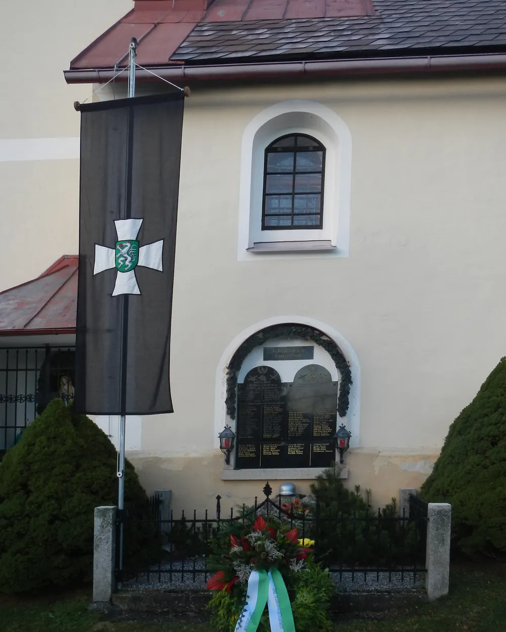 Schwarze Fahne mit weißem Kreuz am Kriegerdenkmal neben der Pfarrkirche, darunter ein Kranz