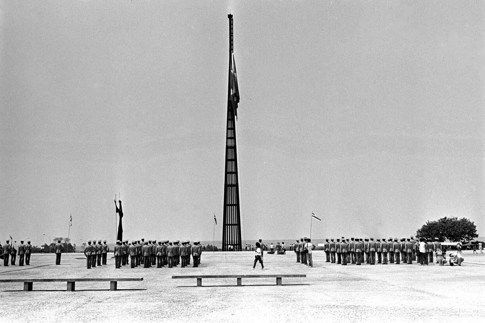 Fotografia em preto e branco do Mastro da Bandeira na Praça dos Três Poderes, em Brasília, com uma tropa em forma