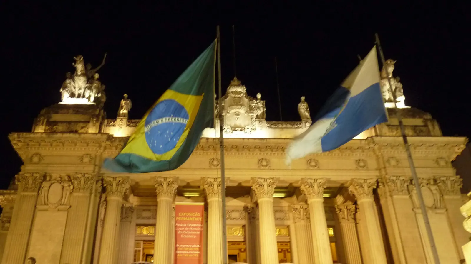 A Bandeira Nacional e a bandeira do Rio de Janeiro iluminadas à noite diante do Palácio Tiradentes