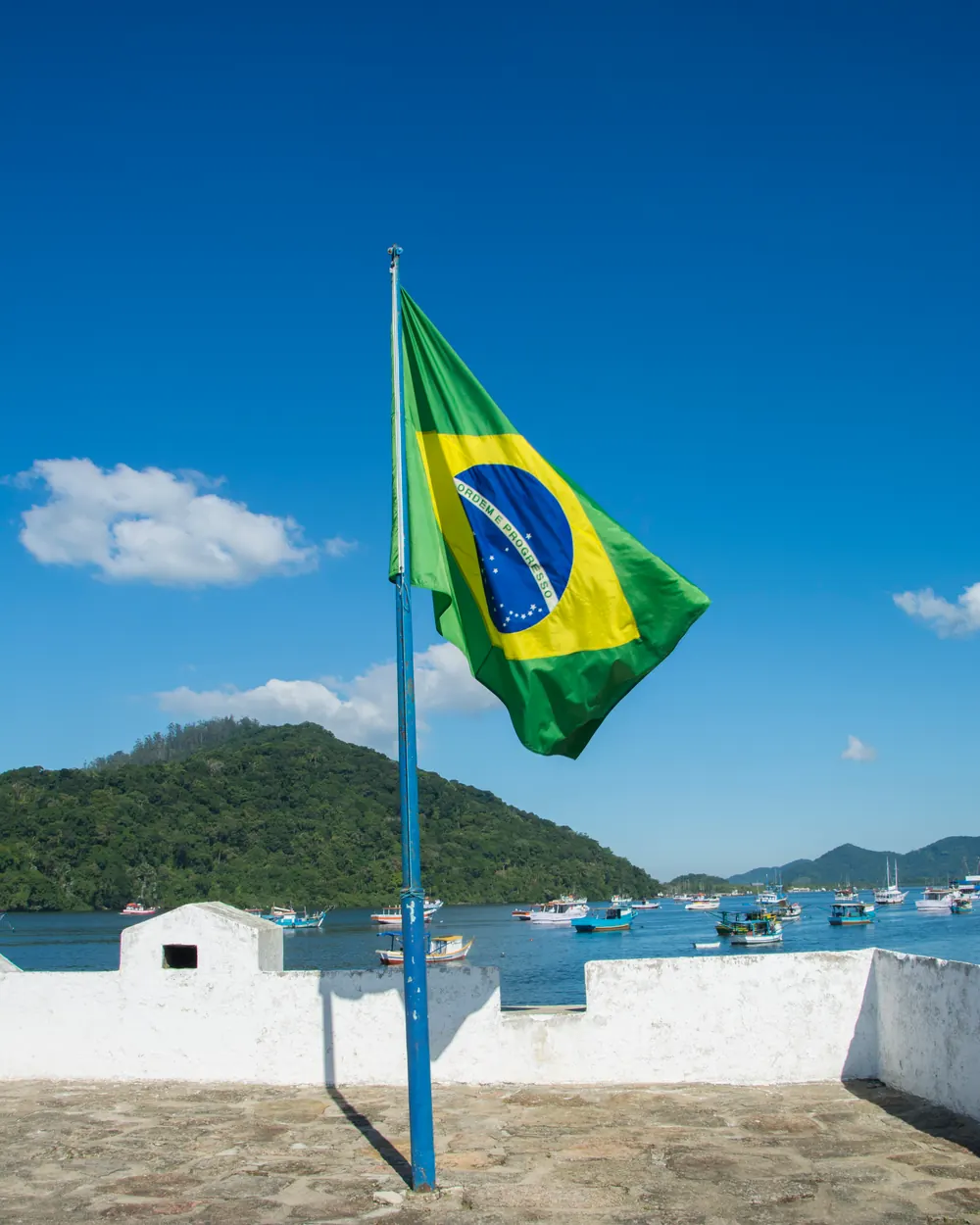 A Bandeira Nacional em um mastro azul sobre o pátio de um forte à beira-mar, com morros verdes ao fundo