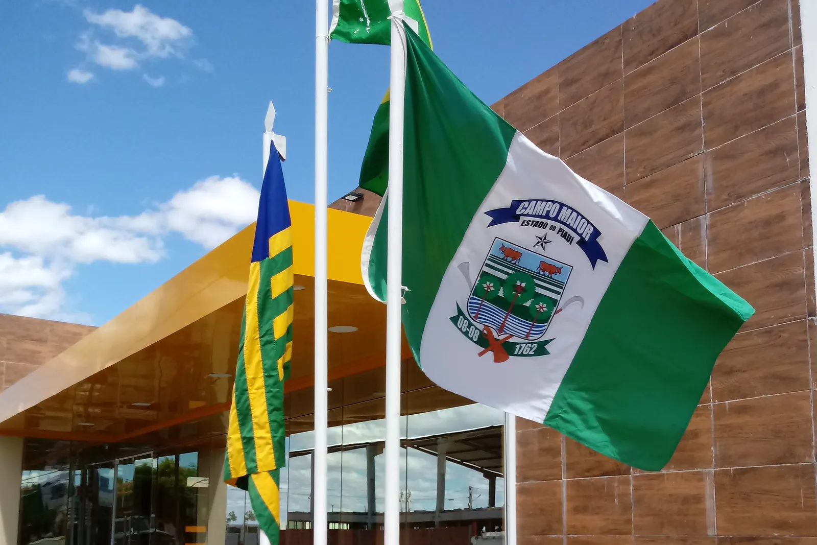 Três mastros com a Bandeira Nacional ao centro, a bandeira do estado e a do município, diante de um prédio público