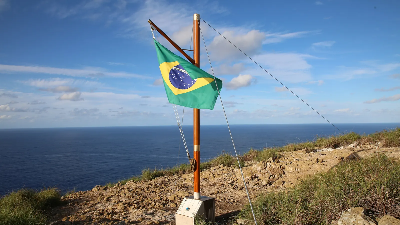 Um mastro de madeira estaiado com a Bandeira Nacional no alto da Ilha da Trindade, com o oceano ao fundo