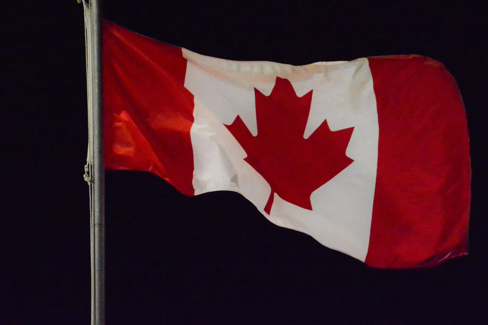 The flag of Canada on a flagpole, lit against the black night sky