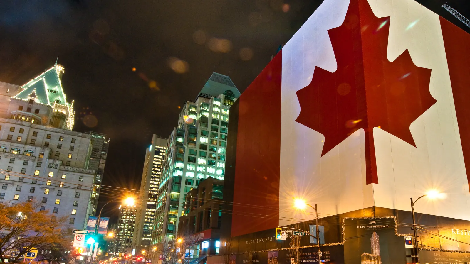 A large flag of Canada lit up at night on a building in Vancouver, with traffic lights streaking past