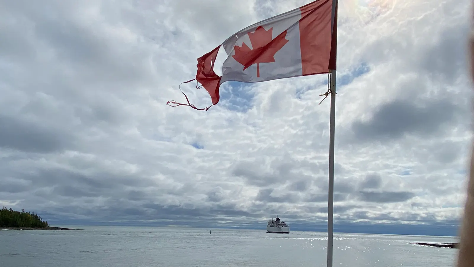 A worn flag of Canada with frayed edges flying in the wind by a lake