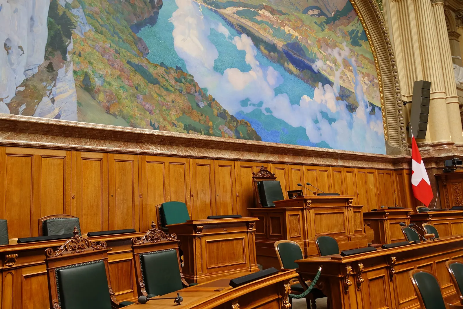 Der Nationalratssaal im Bundeshaus mit Holzpulten, Wandbild und einer Schweizerfahne