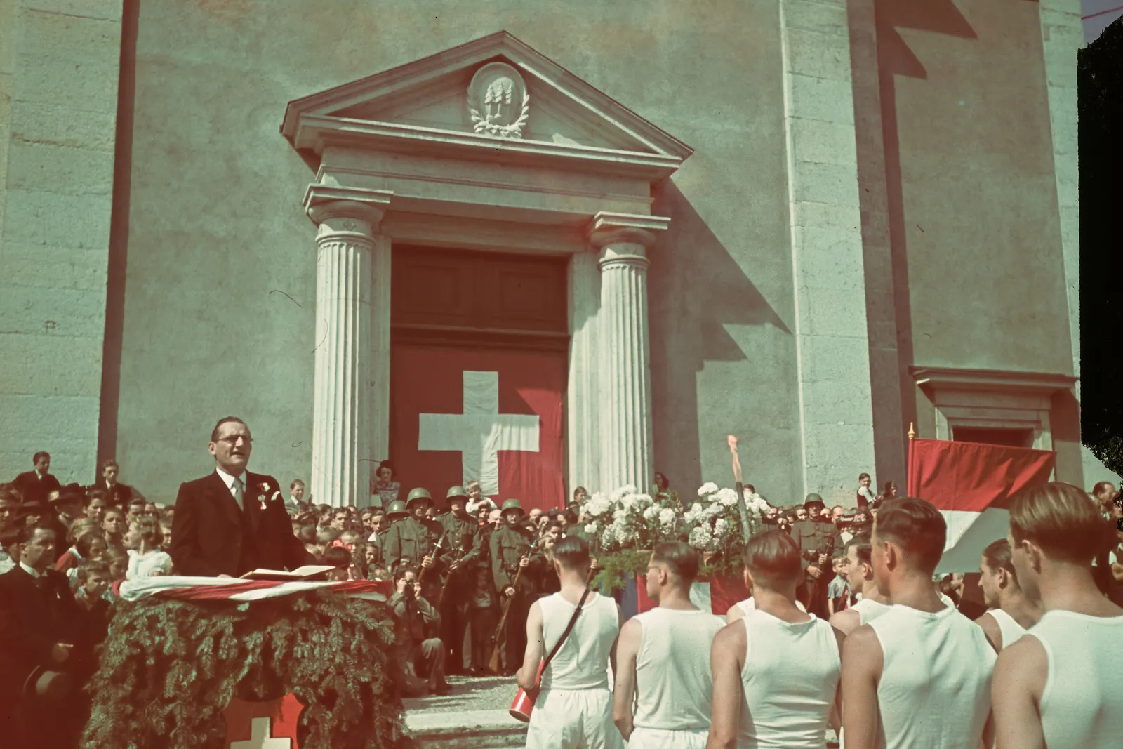 Farbfotografie einer Feier vor einer Kirche mit Schweizerkreuz, ein Redner und junge Turner davor