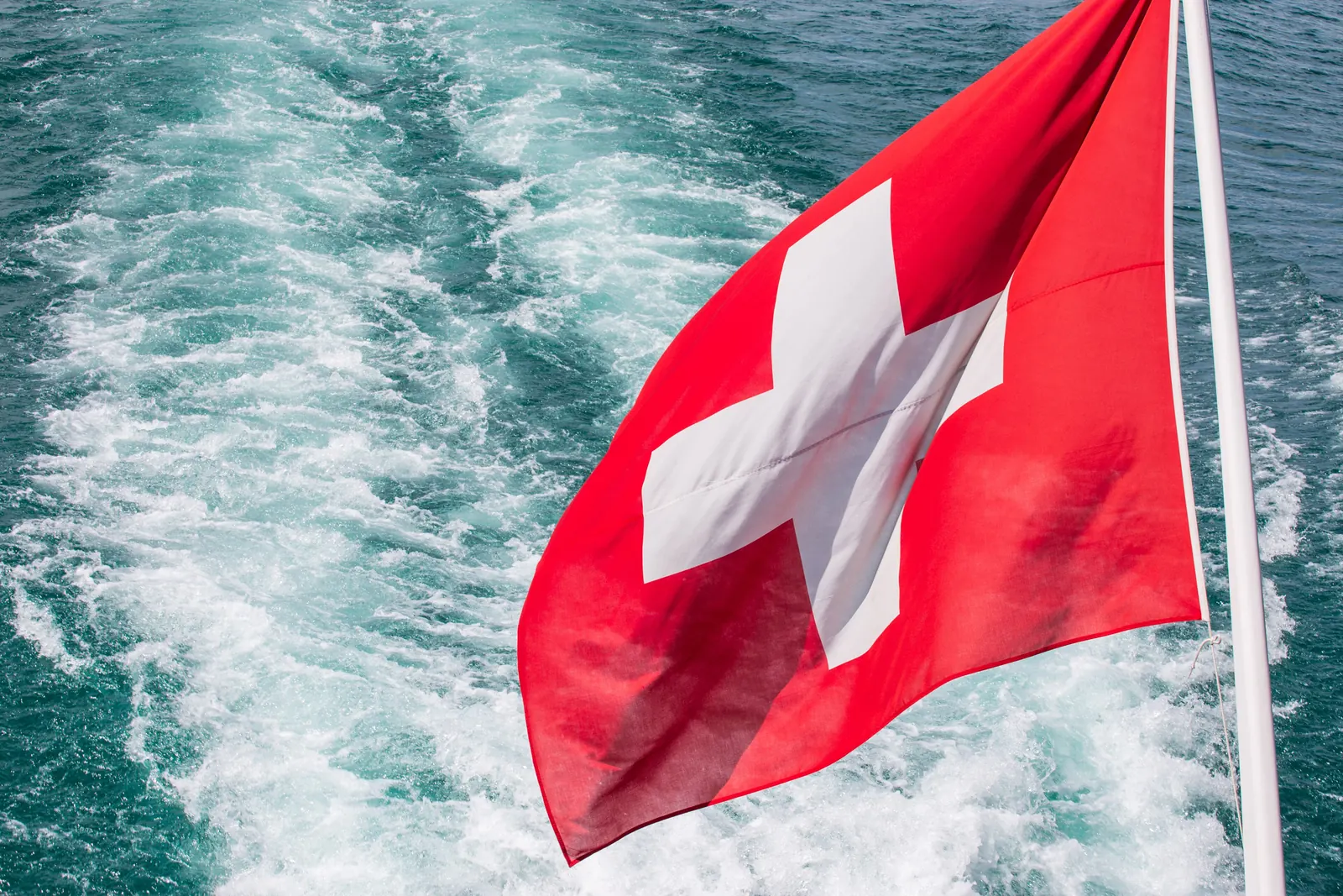 Die quadratische Schweizerfahne am Heck eines Schiffs auf dem Thunersee, dahinter das Kielwasser