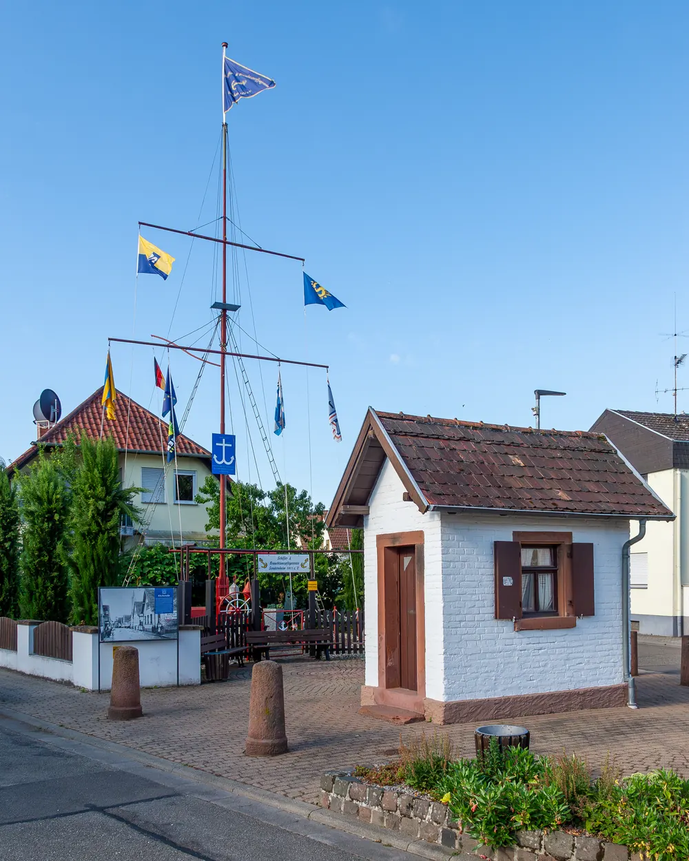 Hoher Fahnenmast mit Querbalken und Flaggen vor einem kleinen Haus in Sondernheim