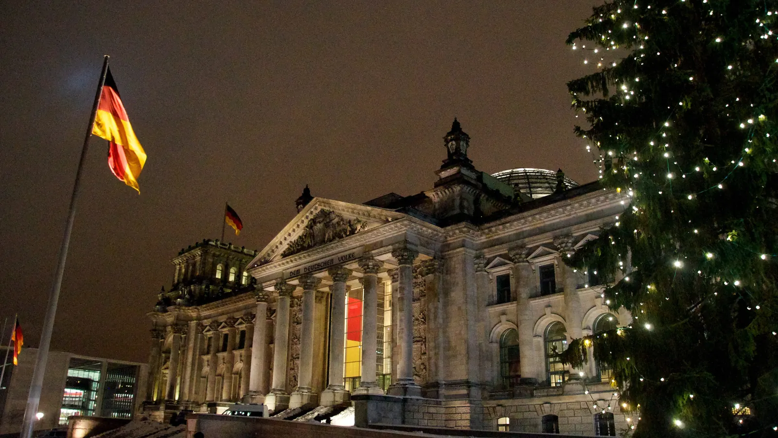 Das angestrahlte Reichstagsgebäude am Abend mit der Bundesflagge am Mast und einem beleuchteten Weihnachtsbaum