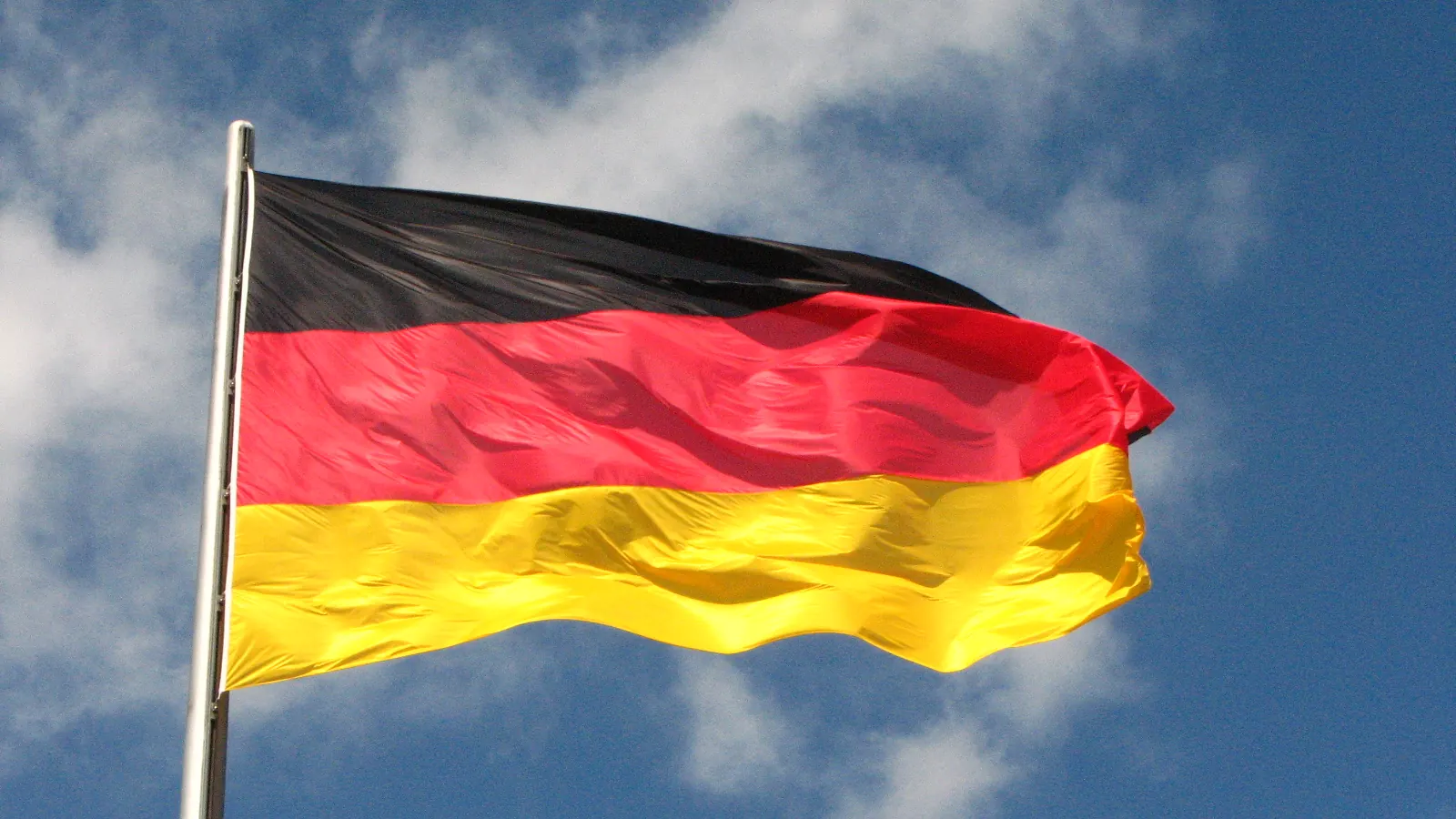 Die deutsche Flagge in Schwarz, Rot und Gold weht an einem Mast vor Wolken und blauem Himmel