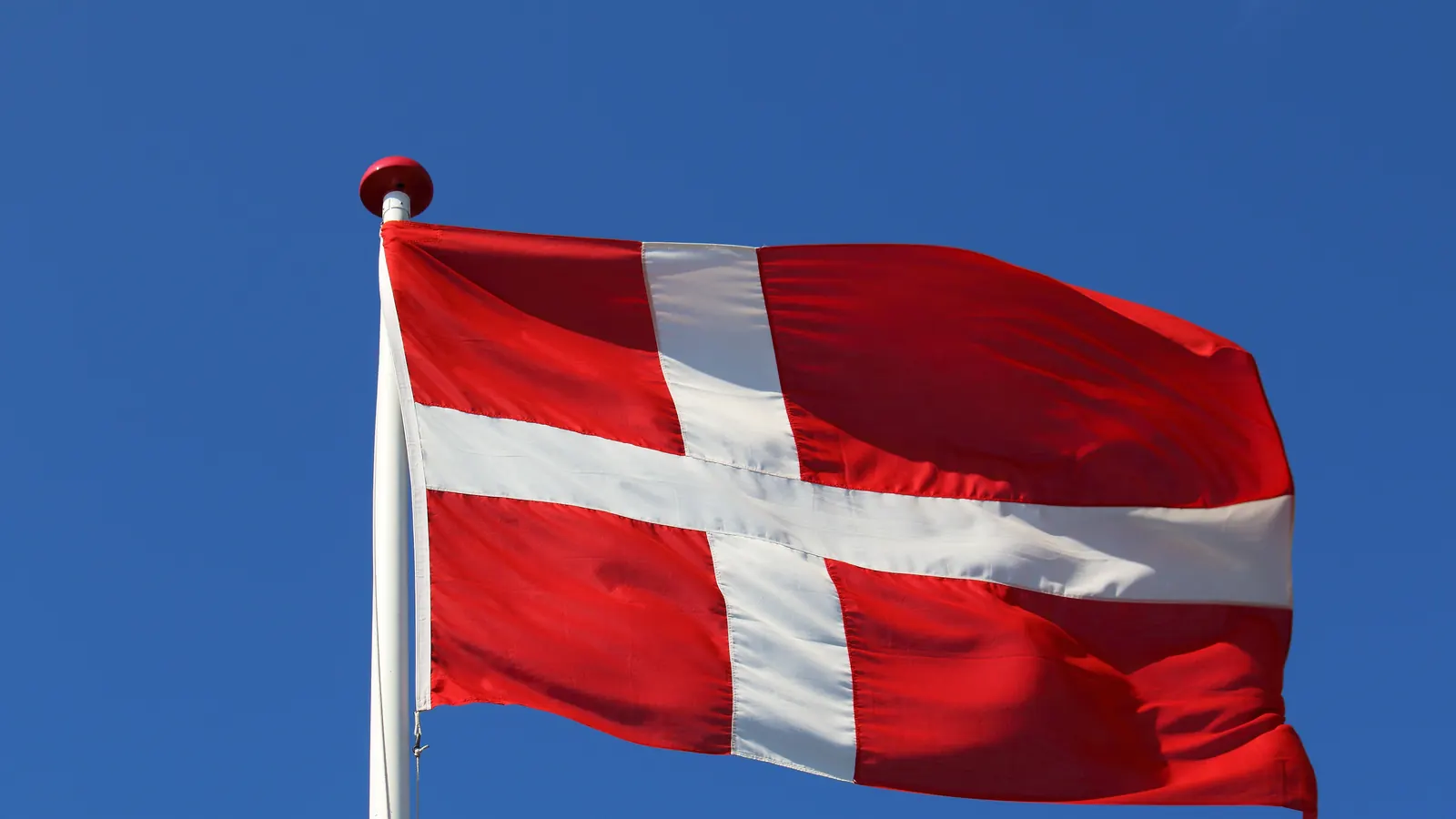 Dannebrog vajer på en hvid flagstang med rød knop mod en klar blå himmel