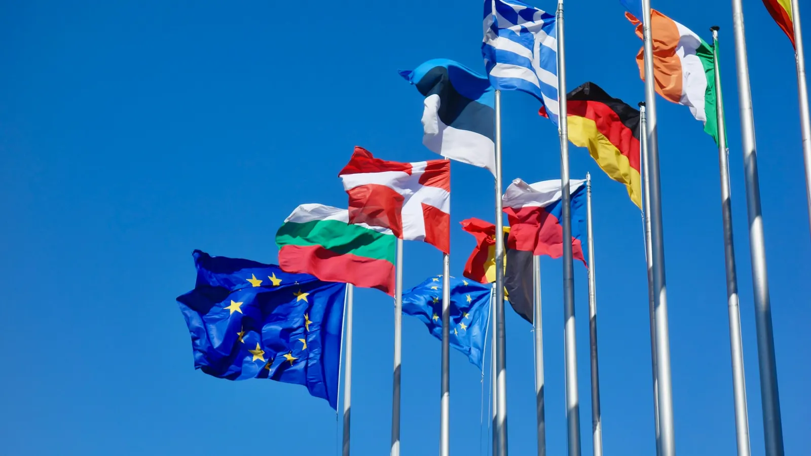 EU-flaget og flag fra flere europæiske lande på en række høje flagstænger mod blå himmel