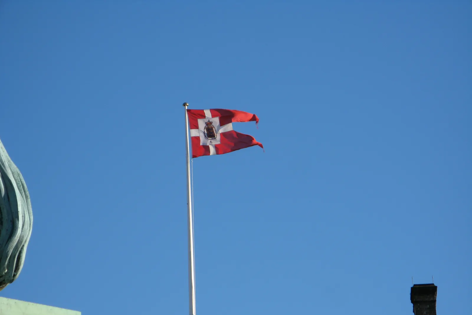 Splitflag med kongeligt våben i et midterfelt på en flagstang mod blå himmel