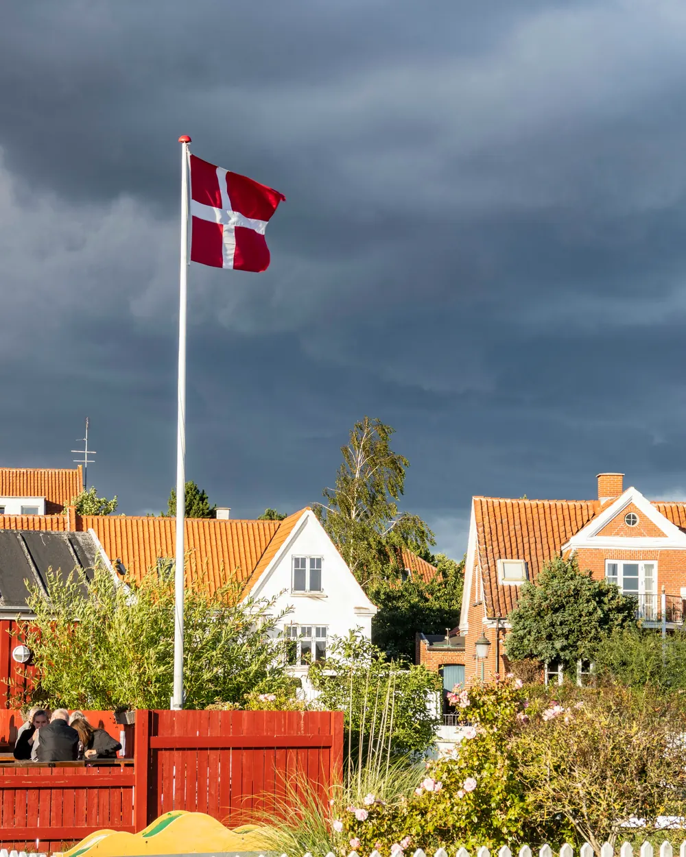 Hvid flagstang med Dannebrog i en have bag et hvidt stakit og røde tegltage