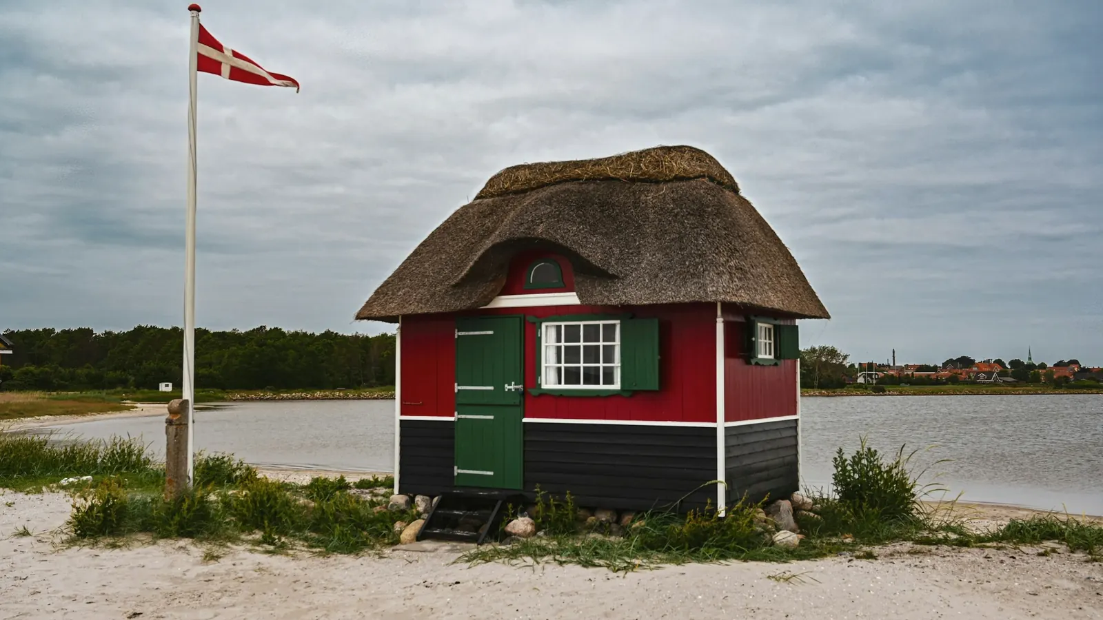 Lille stråtækt badehus på en strand med Dannebrog på en hvid flagstang
