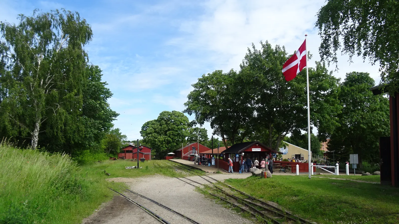 Dannebrog på en hvid fritstående flagstang ved en lille station med smalsporede skinner