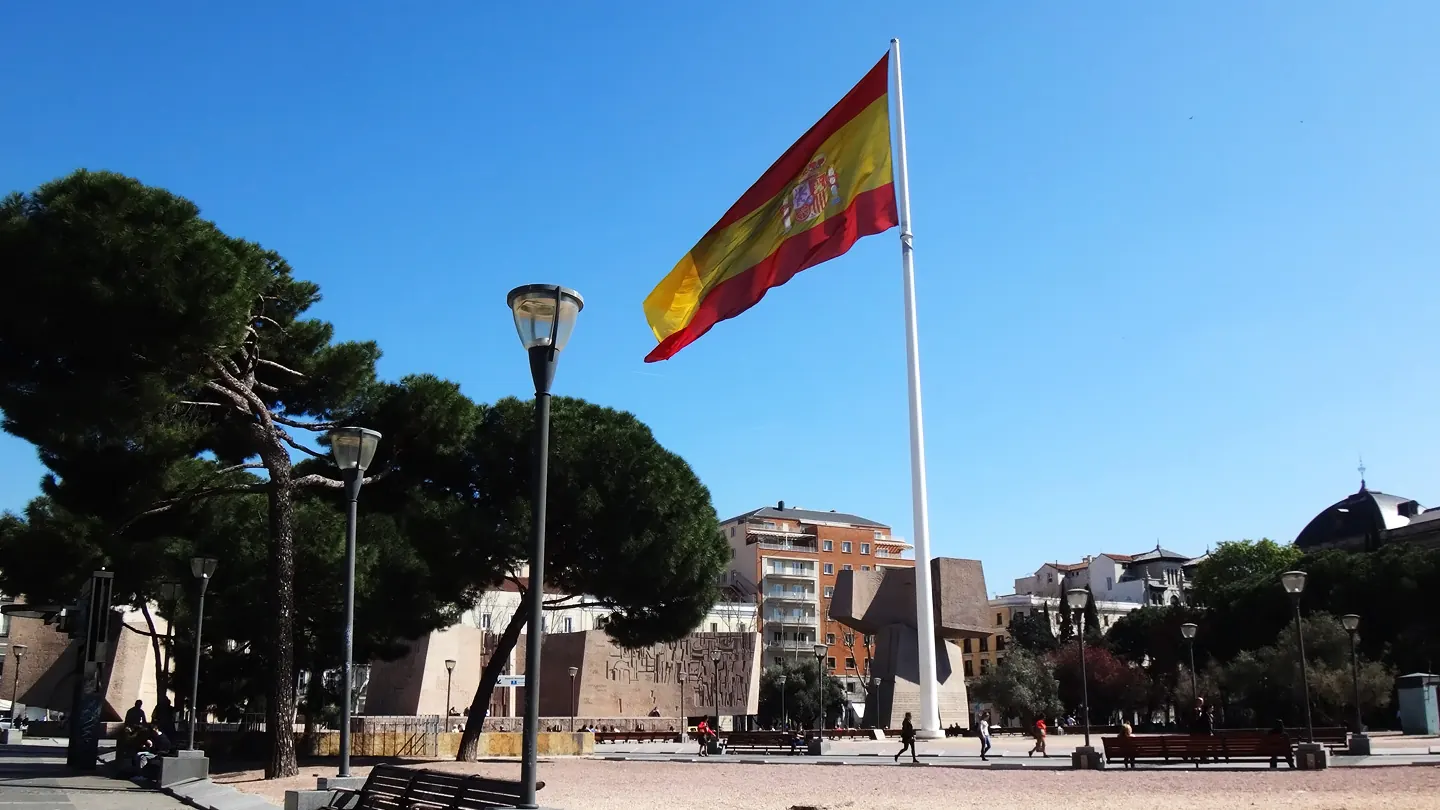 La bandera de España en el mástil de la plaza de Colón de Madrid, entre pinos y farolas