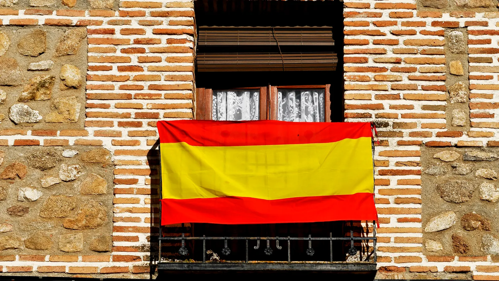 Una bandera de España cuelga de un balcón de hierro en una fachada de ladrillo y piedra