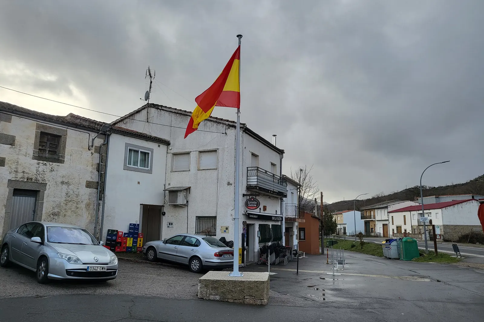 Un mástil exento con la bandera de España en una calle de un pueblo, bajo un cielo nublado