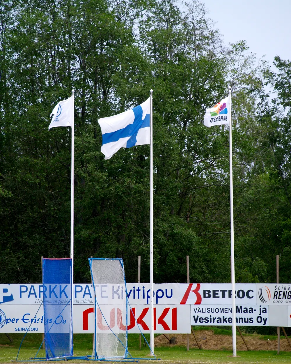 Kolme lippusaloa pesäpallostadionin laidalla, keskimmäisessä Suomen lippu
