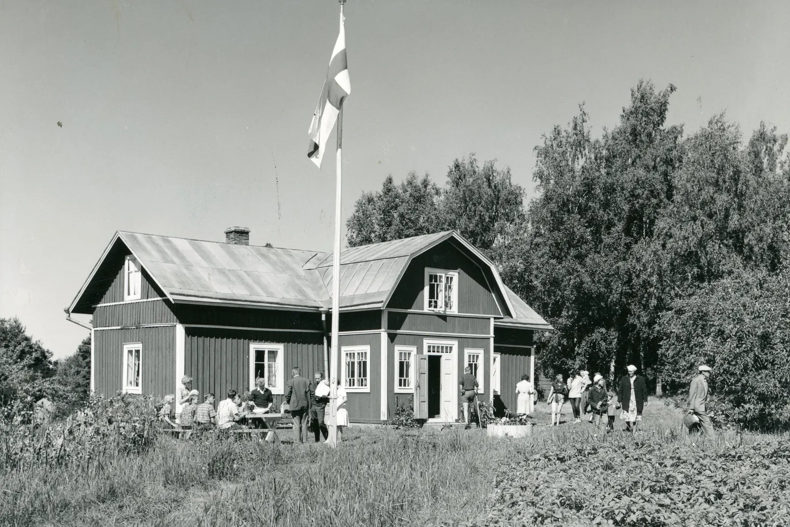 Mustavalkoinen valokuva puutalosta ja sen pihalla olevasta korkeasta lippusalosta