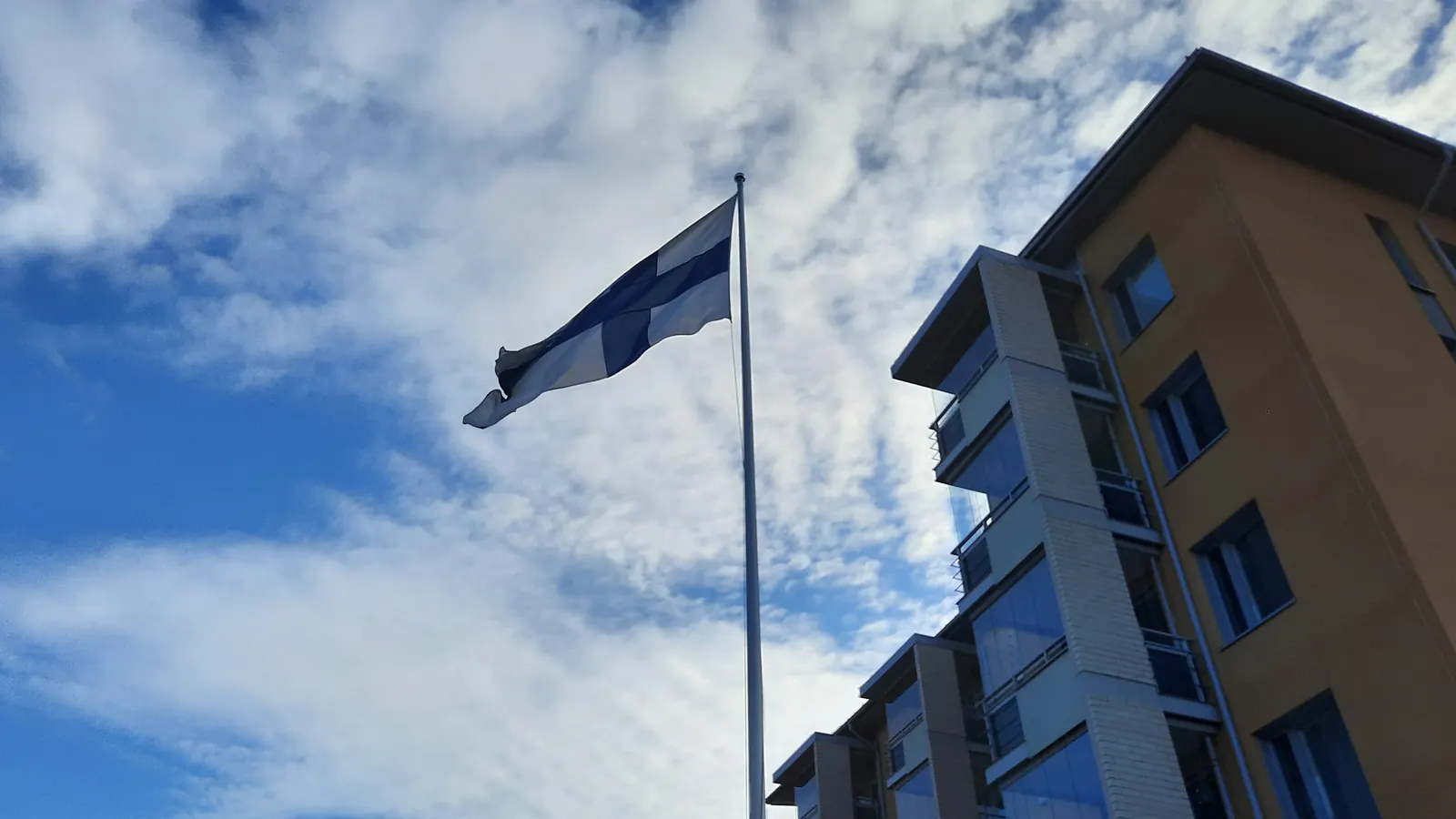 Suomen lippu korkeassa salossa kerrostalon vieressä pilvistä taivasta vasten