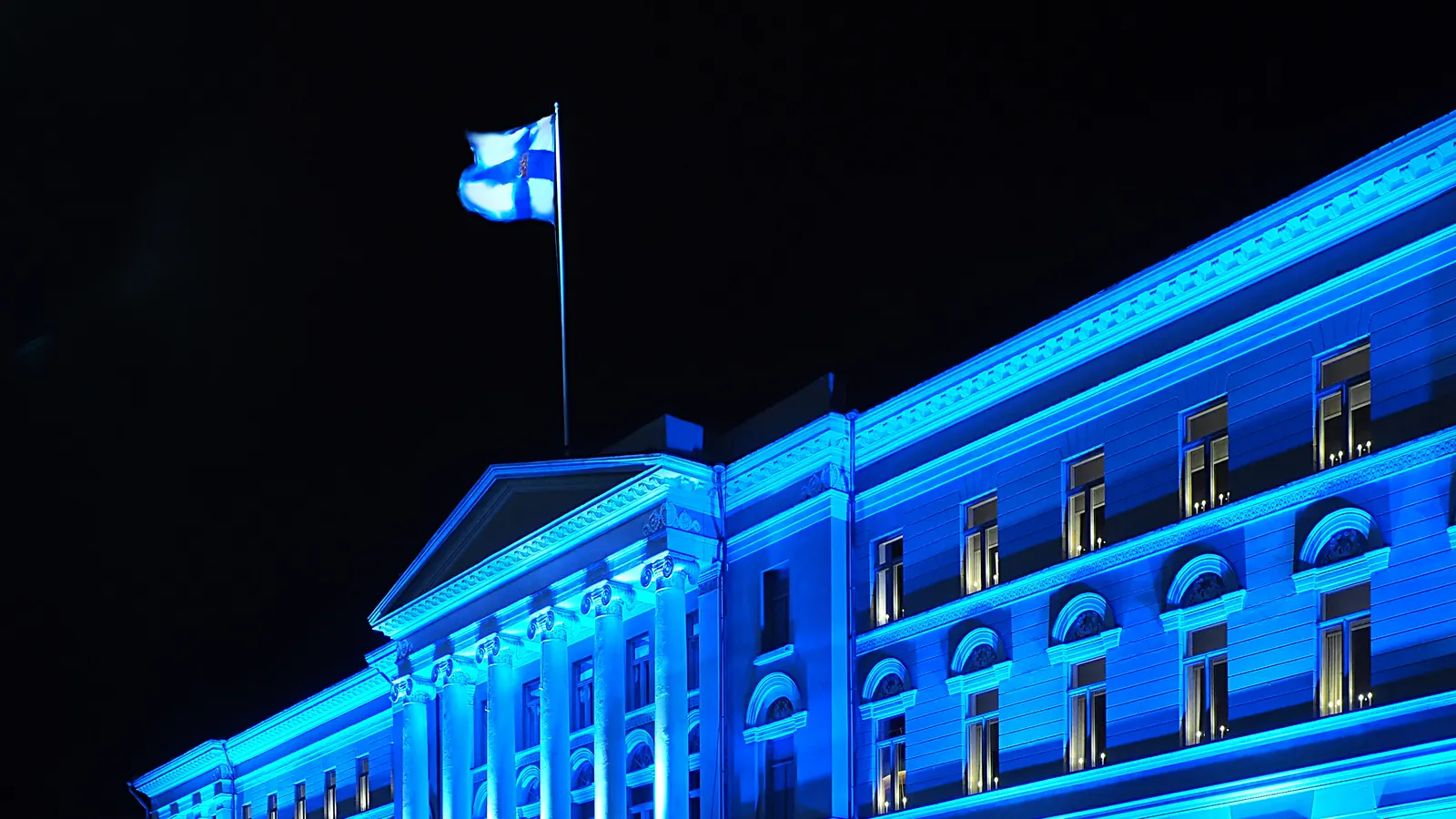Helsingin yliopiston päärakennus valaistuna sinisellä valolla illalla ja Suomen lippu katolla