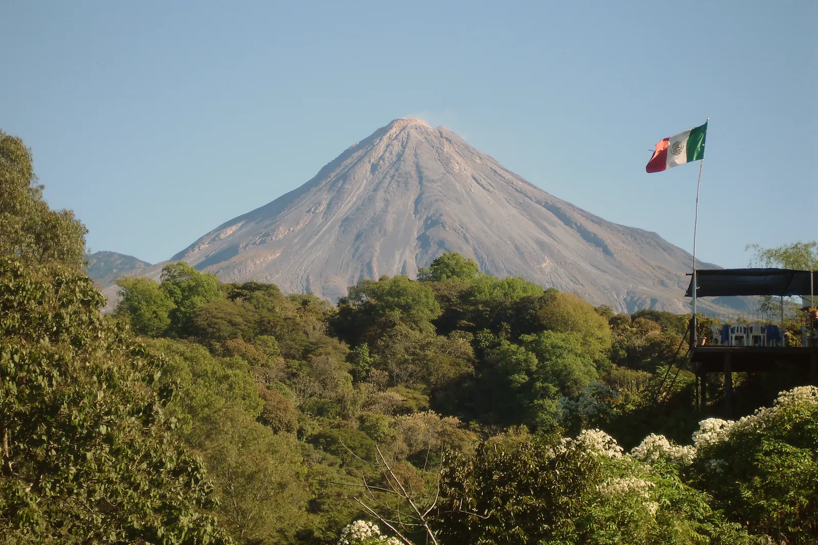 La Bandera Nacional en un asta entre la vegetación, con el Volcán de Colima al fondo