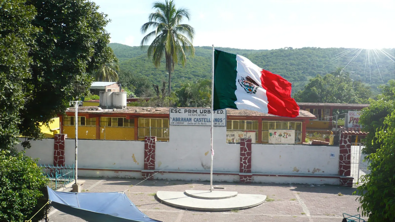 Un asta con la Bandera Nacional en el patio de una escuela, con cerros y palmeras al fondo