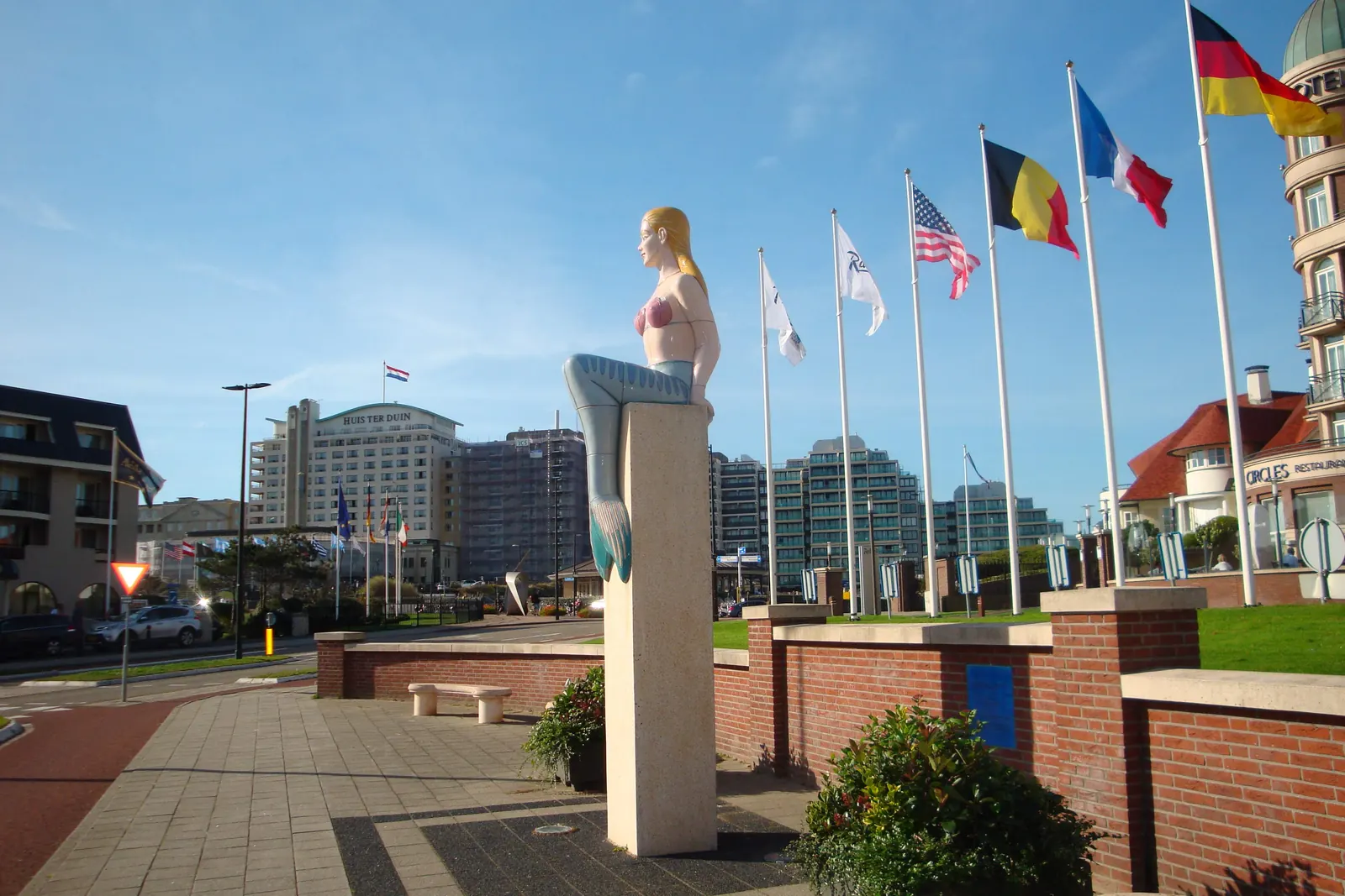 Vlaggen van verschillende landen aan masten bij een beeld van een zeemeermin in Noordwijk