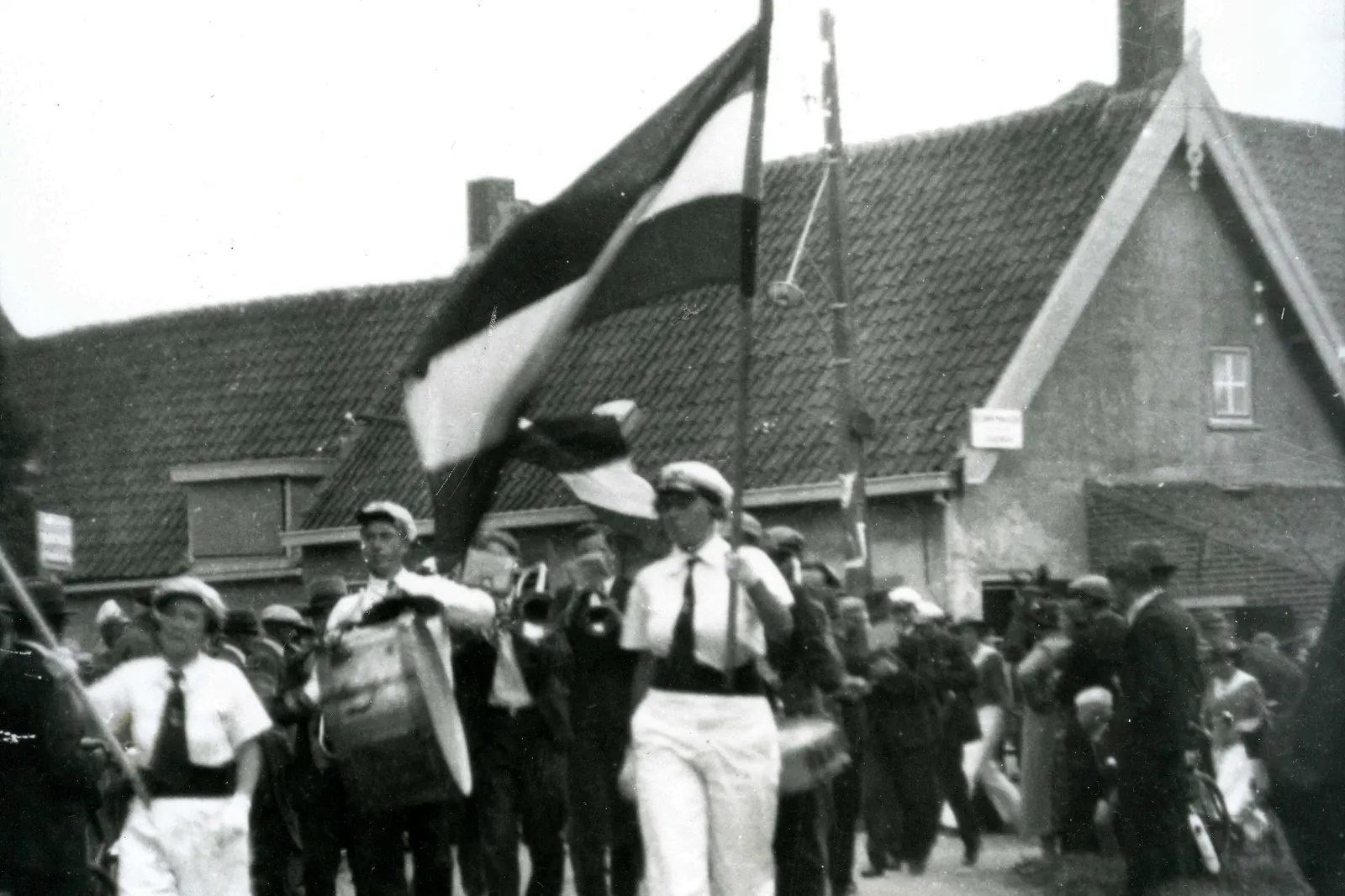 Zwart-witfoto van een fanfare die met de Nederlandse vlag door een straat marcheert