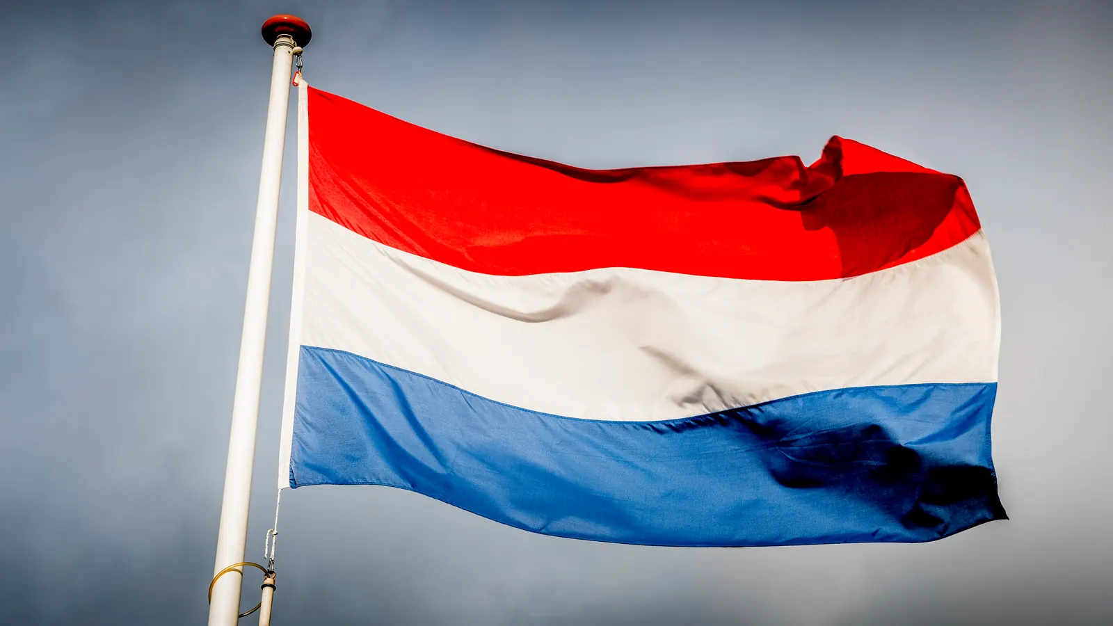 De Nederlandse driekleur in rood, wit en blauw aan een witte mast tegen een donkere lucht