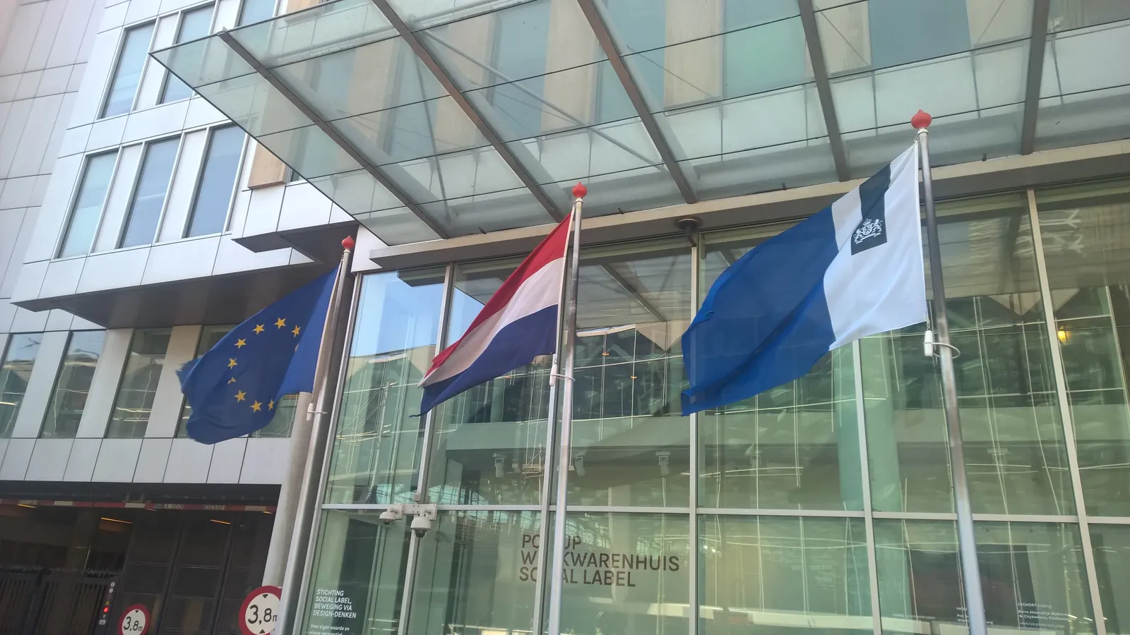 De Europese vlag, de Nederlandse vlag en de vlag van de Rijksoverheid aan masten bij een gebouw in Den Haag, met de Nederlandse vlag in het midden
