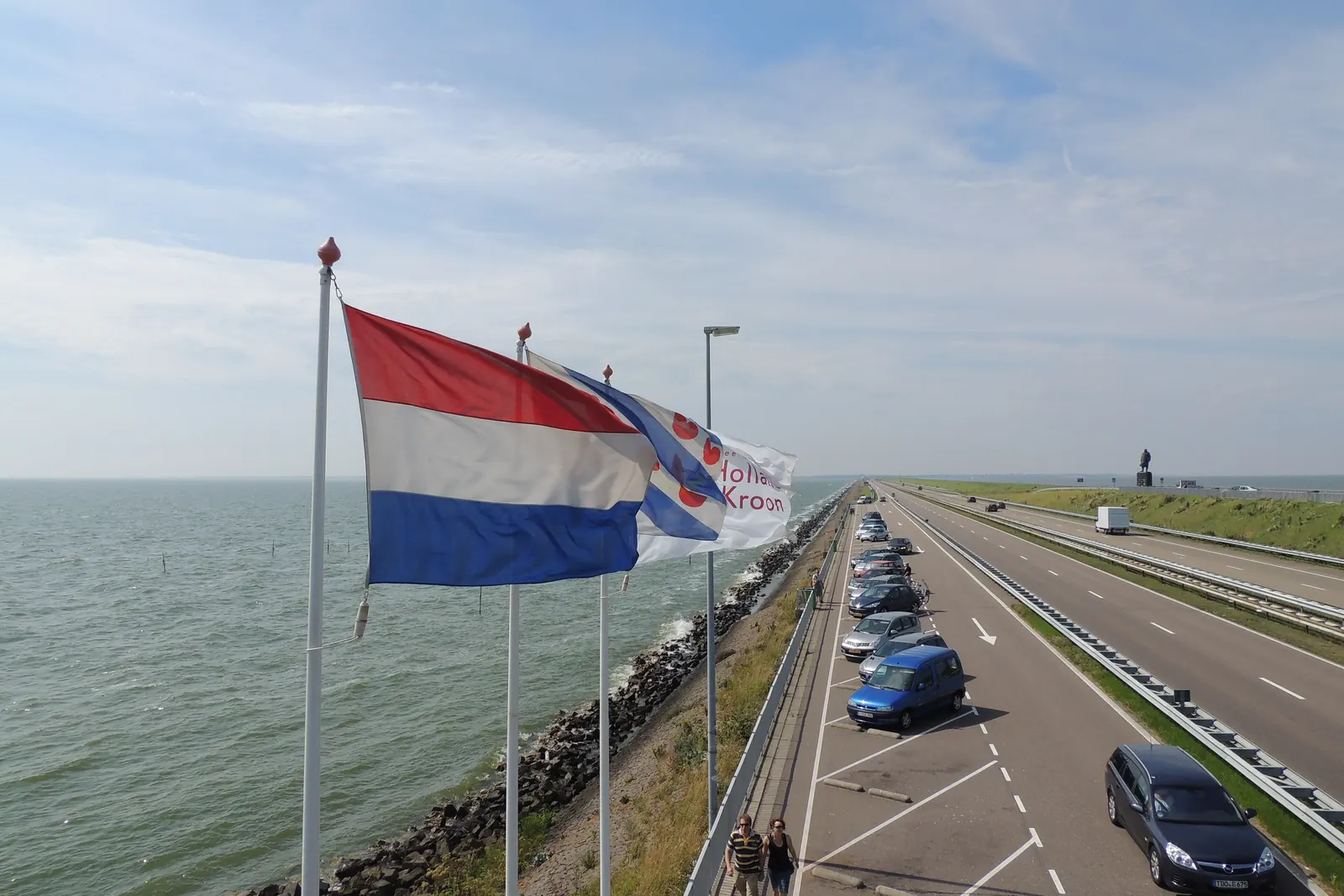 De Nederlandse vlag, de Friese vlag en de vlag van een hotel aan masten op de Afsluitdijk, met de weg en het IJsselmeer