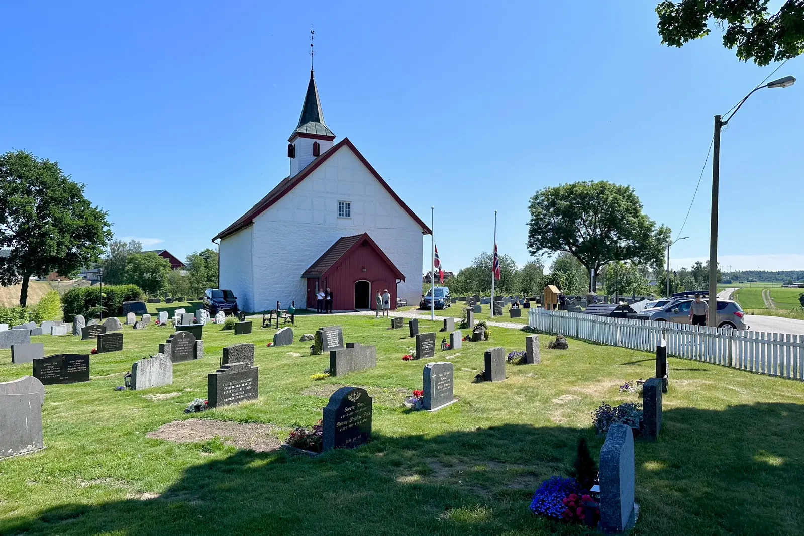Hvit kirke og kirkegård en sommerdag med flagget på halv stang ved Ramnes kirke