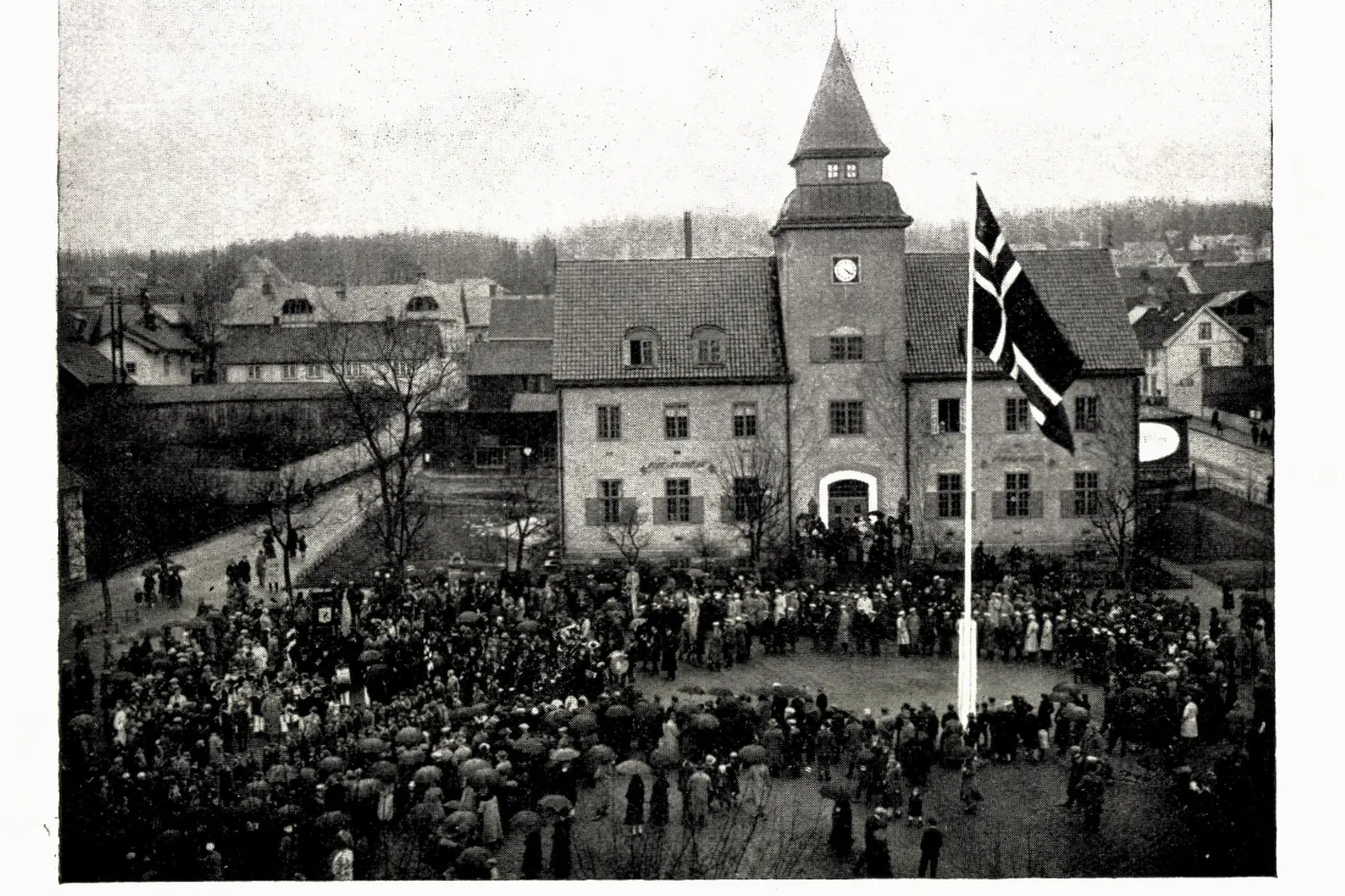 Gammelt fotografi av en folkemengde foran Norges Bank på Lillehammer, med flagget på en høy stang