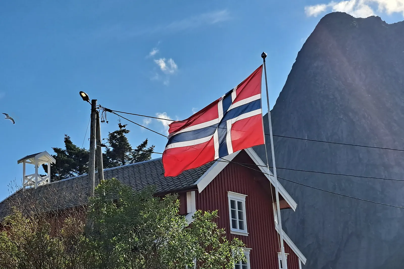 Norsk flagg på en stang ved et rødt hus under en bratt fjelltopp i Reine i Lofoten