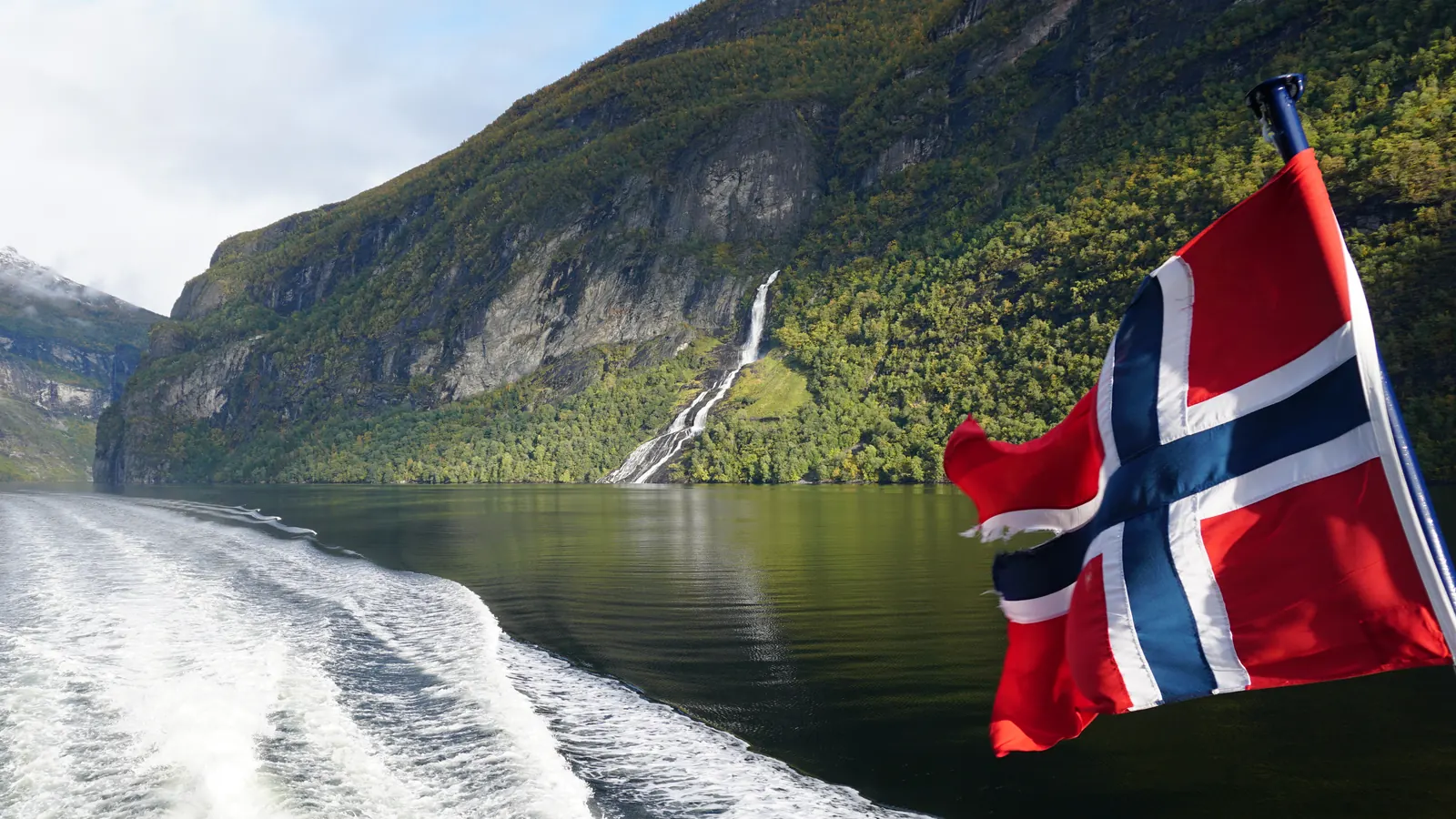 Norsk flagg i akterenden på en båt i Geirangerfjorden, med fjellsider og en foss i bakgrunnen