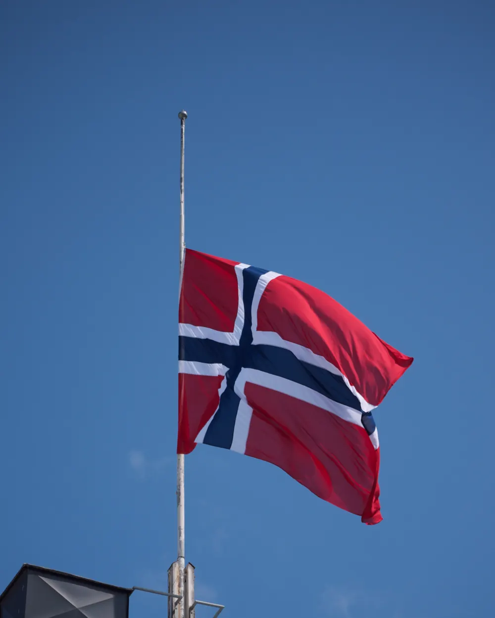Norsk flagg på halv stang på et tak mot blå himmel ved Egertorget i Oslo