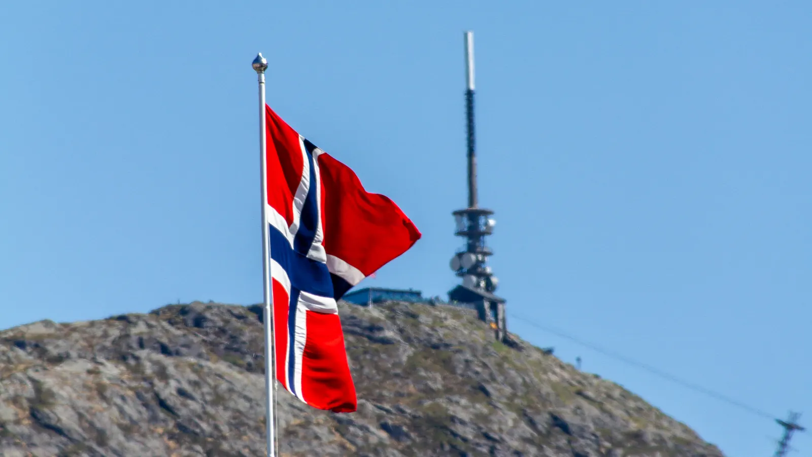Norsk flagg på en hvit flaggstang foran fjellet Ulriken i Bergen