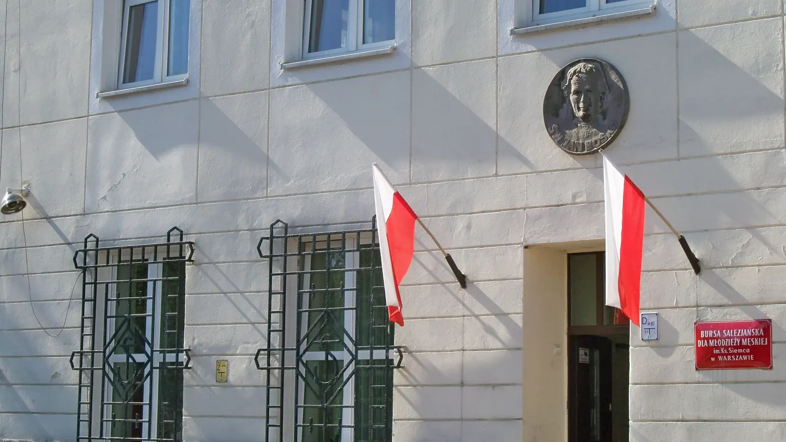 Dwie biało-czerwone flagi na drzewcach w uchwytach przy wejściu do budynku w Warszawie