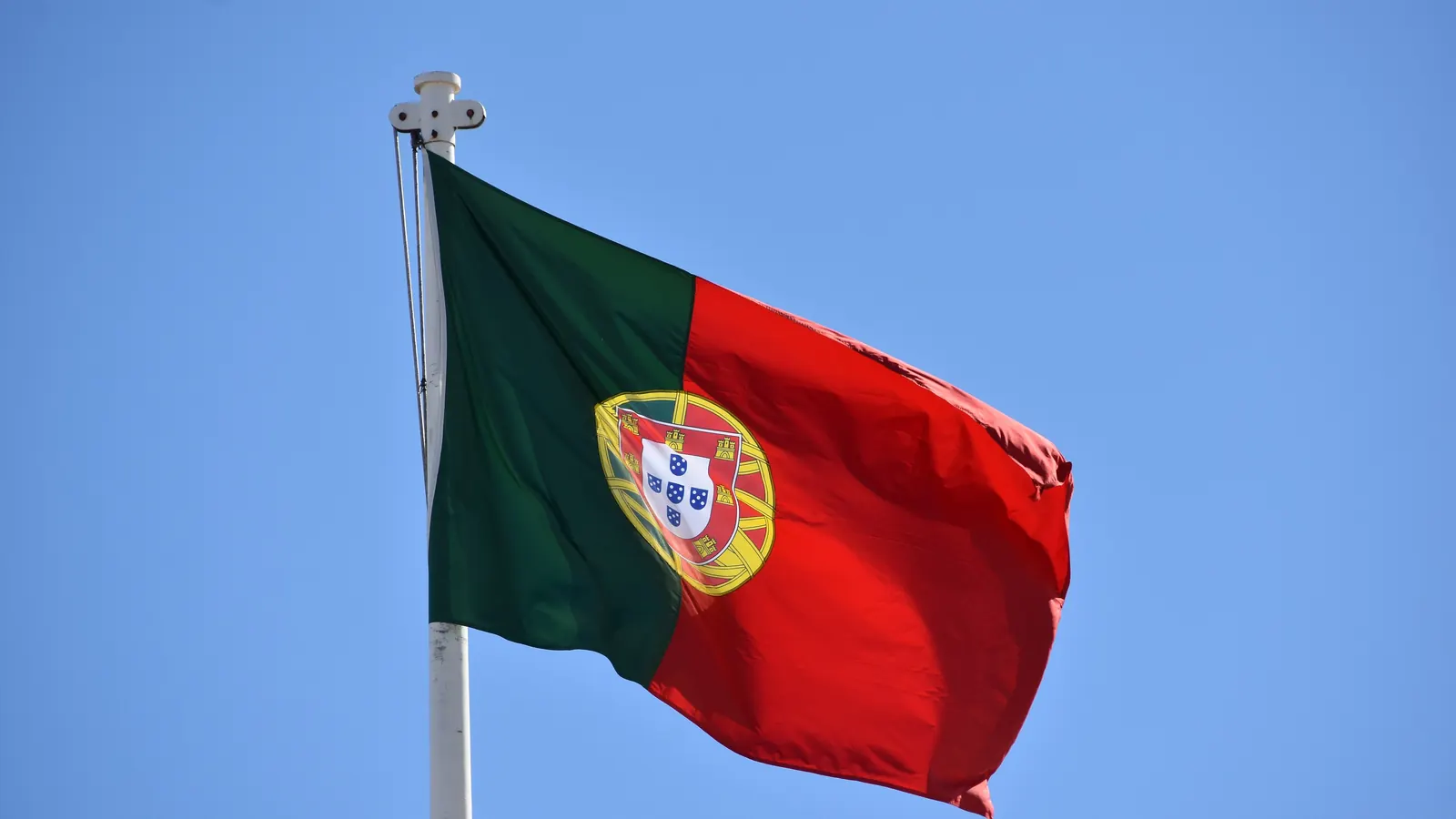 A Bandeira Nacional de Portugal desfraldada no topo de um mastro, contra o céu azul