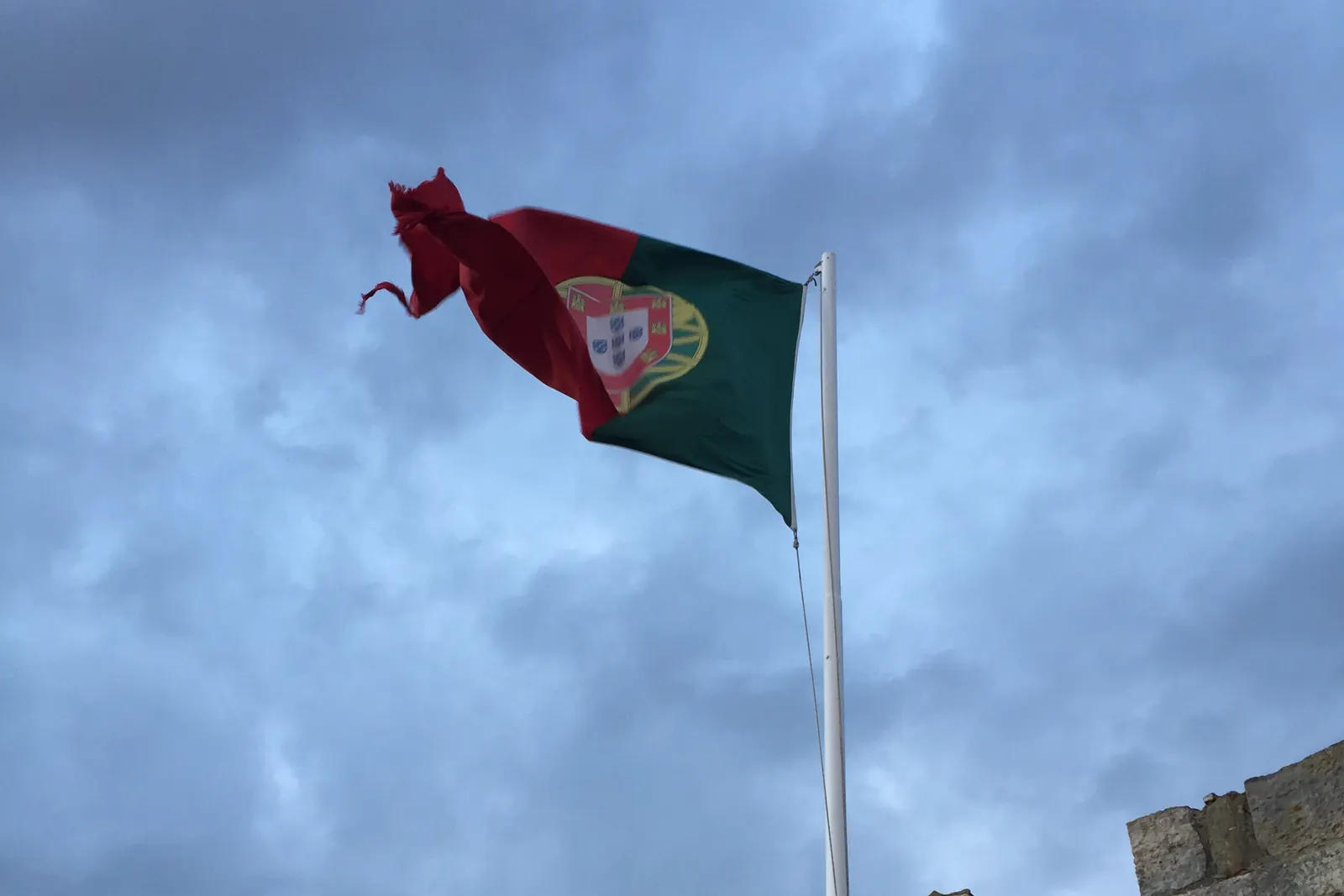 A Bandeira Nacional esticada pelo vento num mastro sobre uma muralha, com nuvens escuras