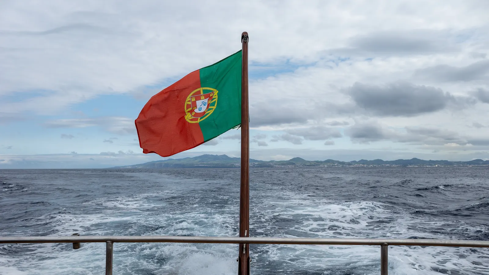 A Bandeira Nacional na popa de um barco de observação de baleias, com o mar e a costa de São Miguel ao fundo