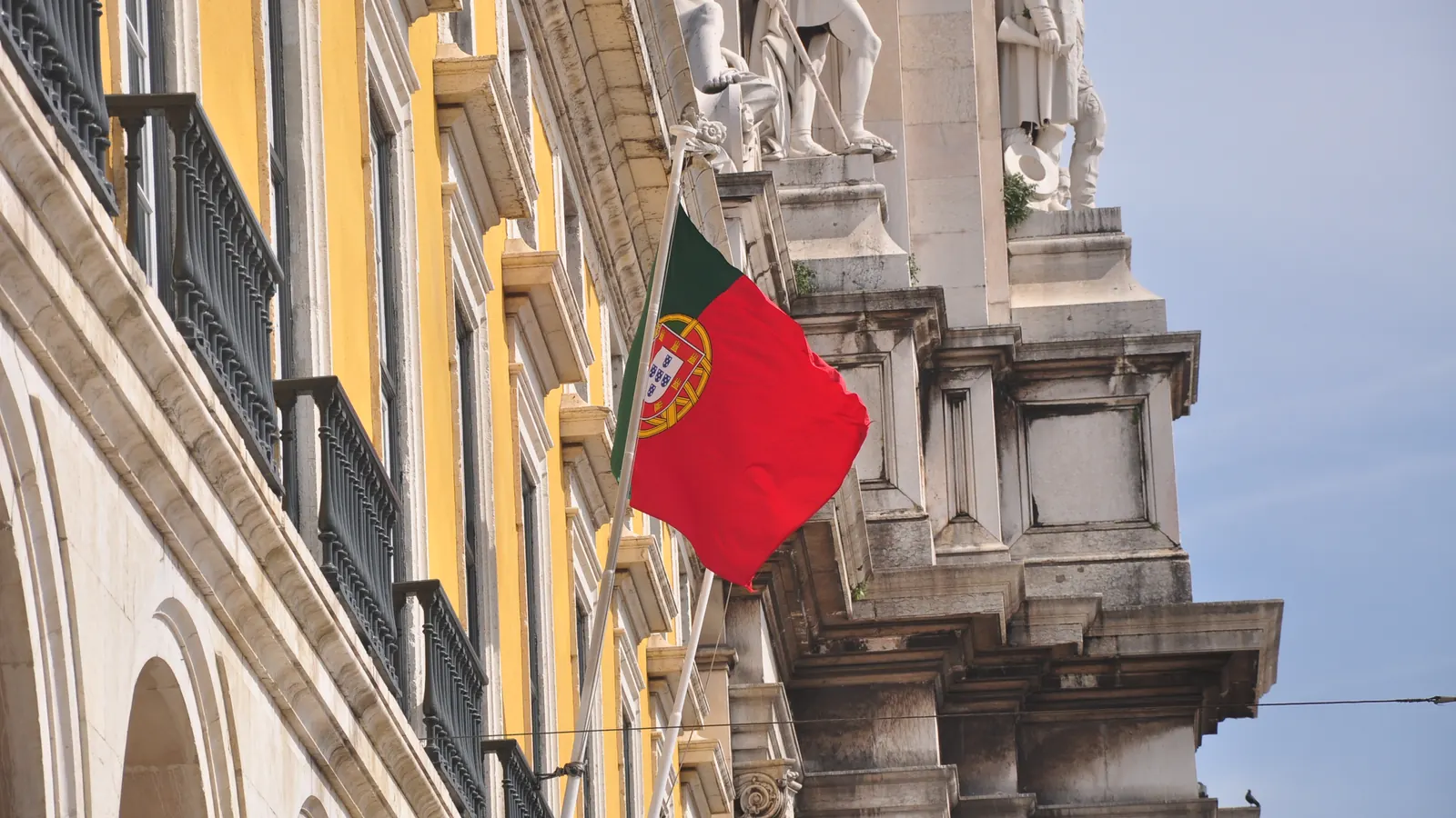 A Bandeira Nacional numa haste de fachada, entre as arcadas e as estátuas de um edifício de Lisboa