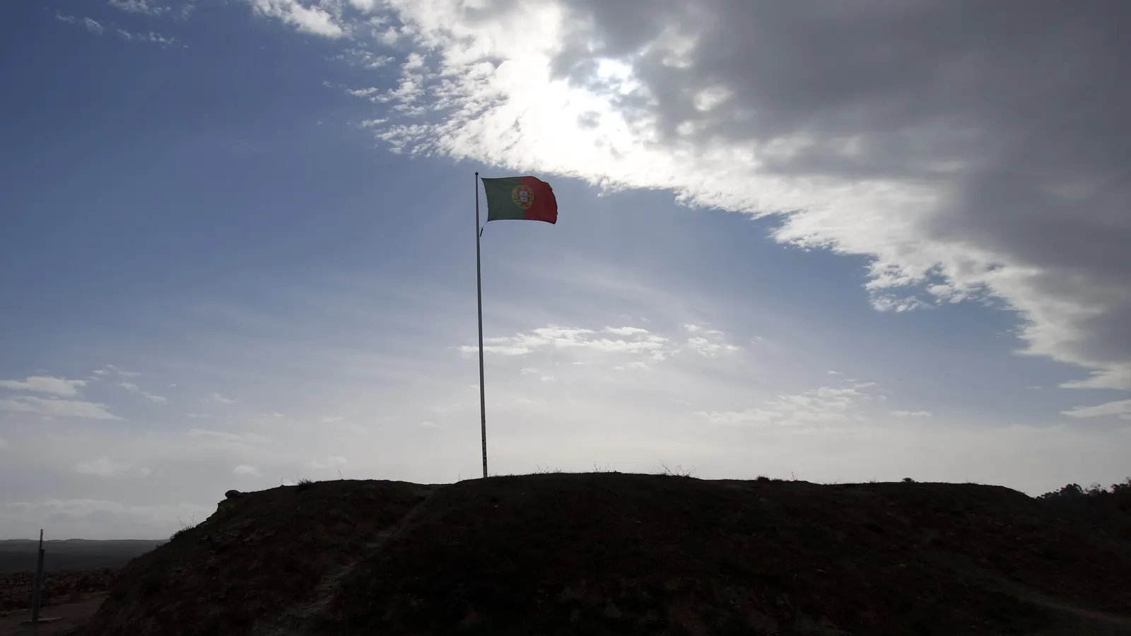 Um mastro com a Bandeira Nacional no alto de uma colina, em contraluz sob um céu nublado