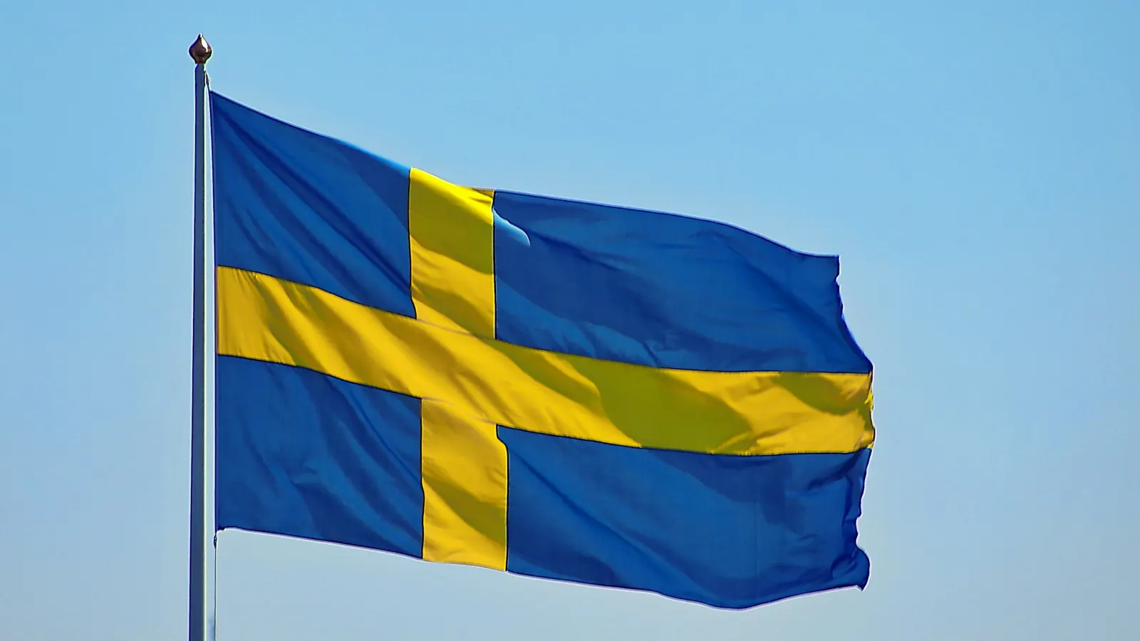 Svensk flagga i blått och gult som vajar på en flaggstång mot ljusblå himmel