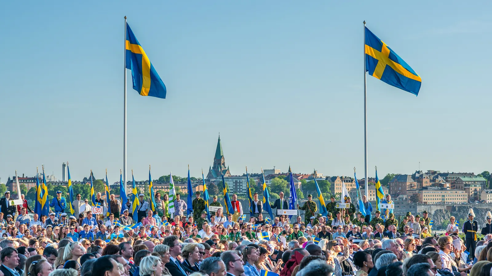 Två svenska flaggor på höga stänger över en stor publik med små flaggor vid ett nationaldagsfirande